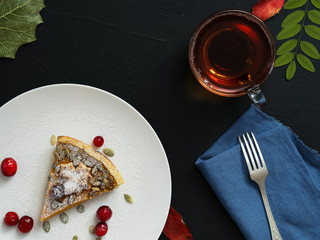 Slice of homemade traditional American pumpkin pie decorated with icing sugar and cranberries on a white plate. Top view. On a black background multi-colored autumn leaves and a cup with tea.