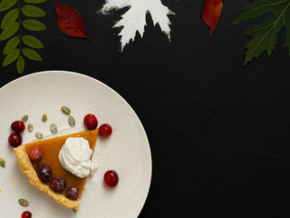 Slice of homemade traditional American pumpkin pie decorated with cream and cranberries on a white plate. Top view. On a black background colorful autumn leaves. Copy space.