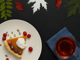 Slice of homemade traditional American pumpkin pie decorated with cream and cranberries on a white plate. Top view. On a black background multi-colored autumn leaves and a cup with tea.
