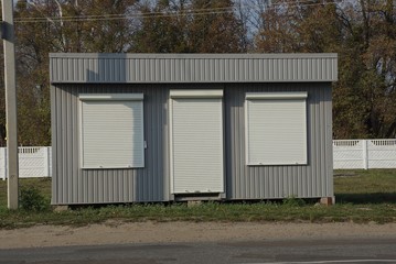 Fototapeta na wymiar gray metal building with windows and a door closed by white roller blinds on green grass by an asphalt road