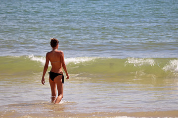 Fototapeta na wymiar Sea vacations, slim girl in bikini going to swim in blue waves, rear view. Concept of swimming, sunny holidays and enjoying the water on paradise coast