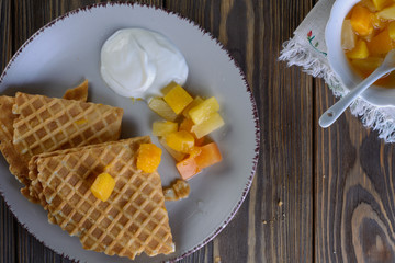 Thin waffles folded as a corner with cream, syrup and slices of fruit on a light ceramic plate on a wooden table ready for breakfast, top view