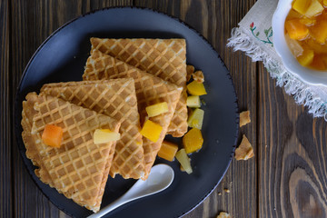 Thin waffles folded as a corner with cream, syrup and slices of fruit on a dark ceramic plate on a wooden table ready for breakfast, top view