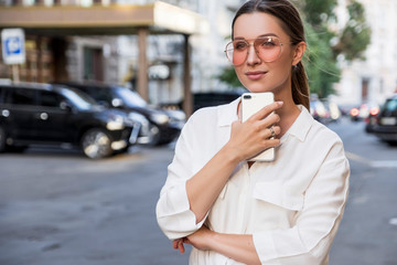 Young caucasian stylish woman in smart-casual outfitand glasses on a european city street. She works on her phone alone outdoors.