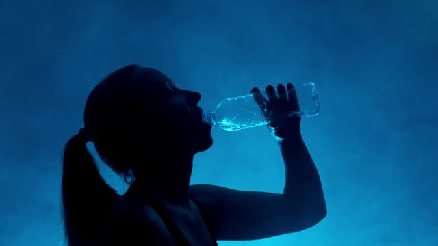 Silhouette of woman drinks water from transparent bottle on dark background with blue smoke. Portrait of woman quenches thirst while taking sip of water from plastic bottle.