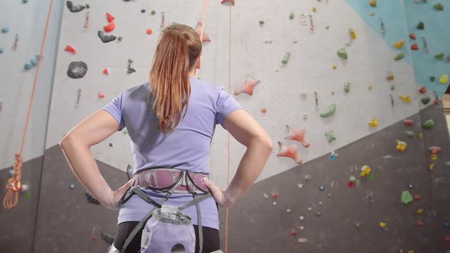 Back view of young Caucasian sporty woman wearing climbing belay standing and looking at climbing wall in gym