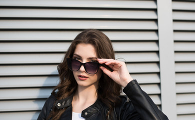 Portrait of european young beautiful smiling woman with dark straight hair in black leather jacket and black sunglasses on horizontal shutters background