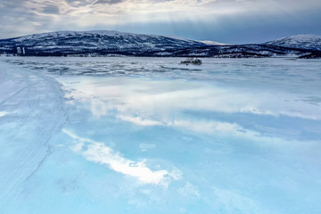 Obraz premium Frozen lake with small island in Northern Mountains starting to melt and water flows on the ice surface. Blue skies and sun rays light down. Joesjo lake in Lappland, Northern Sweden.