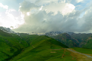 Landscape mountain view peaks in snow and green hills, deep blue sky and huge white clouds background, Caucasian mountains, Kazbek mountain