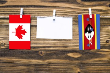 Hanging flags of Canada and Swaziland attached to rope with clothes pins with copy space on white note paper on wooden background.Diplomatic relations between countries.