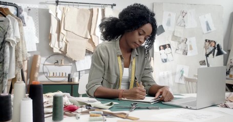 Young pretty African American woman fashion designer working at the laptop computer and drawing sketches in her notebook inthe studio.