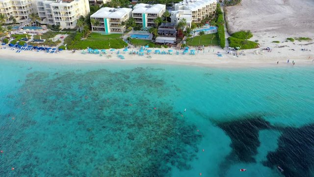 Aerial: Beachfront Resorts, Swimming Pools and People Lounging Under Bright Blue Umbrellas on Beach - Providenciales, Turks and Caicos