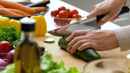 healthy eating, vegetarian food and cooking concept - young woman with kitchen knife chopping cucumber on wooden cutting board at home - Powered by Adobe