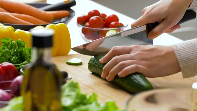 healthy eating, vegetarian food and cooking concept - young woman with kitchen knife chopping cucumber on wooden cutting board at home