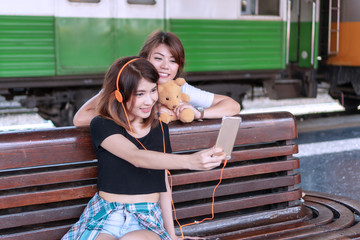 Traveler two young women using smartphone selfie picture to social media in the station. Cheerful girl wear orange earphone with her friend taking photo together while wait train. Friendship concept.