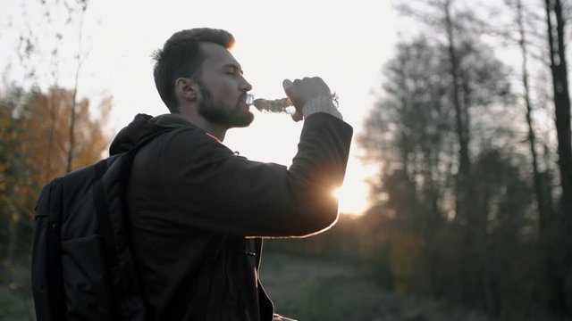 Portrait of young bearded man drinking water from bottle in sunset sun light standing in meadow by autumn forest. Male traveler hiker with backpack relaxing in evening after trekking