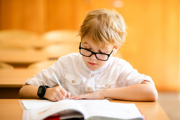 Naklejka na ściany i meble Seven years old child with glasses writing his homework at school. Boy studing at table on class background