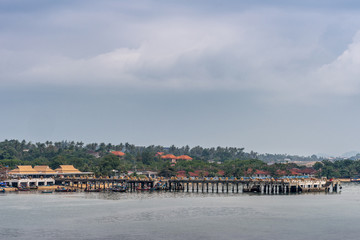 Ko Samui Island, Thailand - March 18, 2019: Off Wat Phra Yai Buddhist Temple on Ko Phan. Bangrak Seatran Pier with forested background and planty of red and orange roofs under cloudscape.