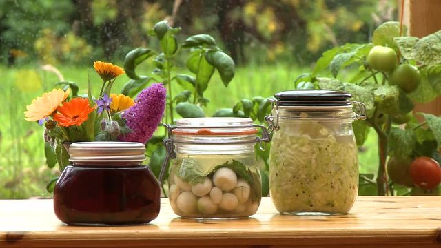 Steady, medium close up shot of jars of garlic cloves, jam and coleslaw on a wooden table.