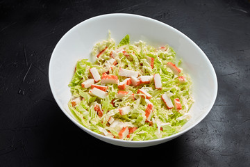 Chopped crab sticks and fresh chinese cabbage leaves in white ceramic bowl on black stone background. Vegetable and imitation crab meat, sea food.  Cooking salad