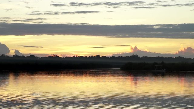 Glowing sunset over calm river, small boat moving slow in distance, wide shot