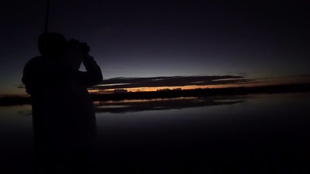 Silhouette of person looking through binoculars over lake at dusk, last light on horizon