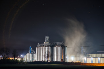 Grain dryer plant working at night.  Floating steam around. Agro farm metal elevator, tall silos with dark  sky on the background. © mckornik