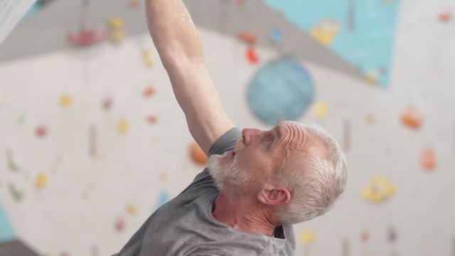 Close up of elderly Caucasian sporty man with grey hair and beard wearing safety equipment practicing bouldering on artificial rock