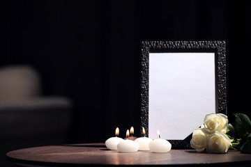 Funeral photo frame with black ribbon, roses and candles on wooden table indoors. Space for design