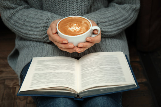 Woman with cup of coffee reading book at home, closeup