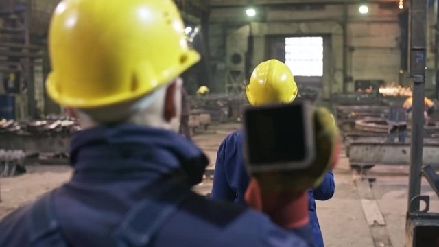Dolly shot with rear view of workers in uniforms and hard hats carrying hollow steel beam through metal fabrication facility