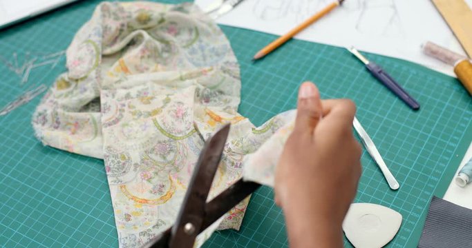 View ver the shoulder on the African American female clothing designer working at the desk and cutting the fabric. Close up.