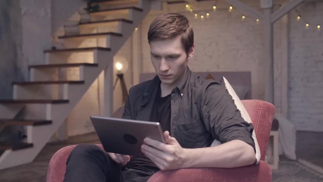 Modern decorated home interior on the background, professional photo and video shooting, close-up. Stock footage. Young man sitting in comfortable armchair and looking at digital tablet