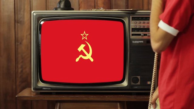Young Man Talking on Old Phone at Home and Watching a Red Soviet Union Flag on a Retro TV. 