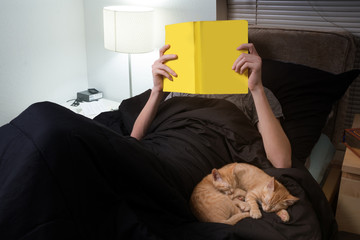 A teenager lies in bed, reads a book, kittens are sleeping nearby, a reading lamp is on.