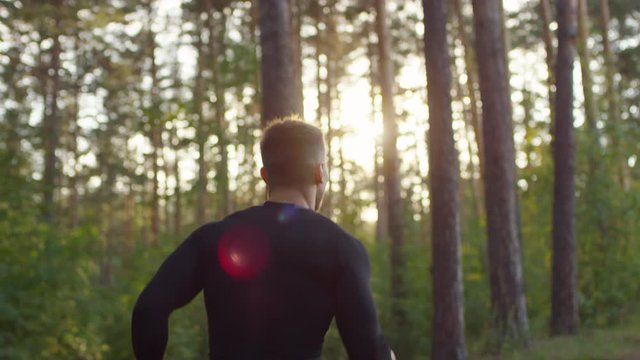 Following shot of muscular handsome man in sportswear running through forest in sunny morning and then turning at camera, smiling and showing V sign