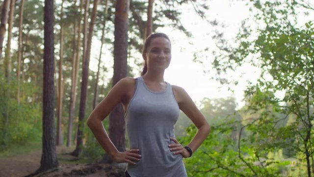 Beautiful athletic woman running towards the camera, tapping on screen of smartwatches and then standing with hands on hips and smiling at camera while training in forest in the morning
