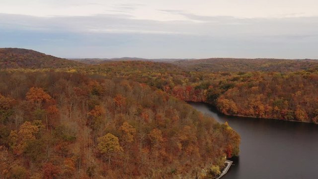 aerial dolly out over the orange colored tree tops & mountains. The drone camera flies on a cloudy day over the falls & dam wall at the New Croton Dam in Westchester County, NY.