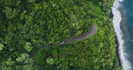 Aerial view of a white luxury car driving on a windy coastal green jungle road with blue ocean, island vacation adventure
