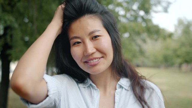 Close-up portrait of young Asian woman looking at camera smiling touching hair standing alone in park. Nature, beautiful people and summertime concept.