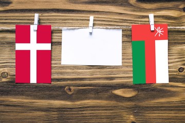 Hanging flags of Denmark and Oman attached to rope with clothes pins with copy space on white note paper on wooden background.Diplomatic relations between countries.