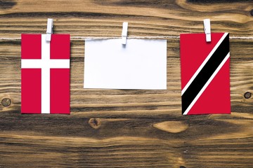 Hanging flags of Denmark and Trinidad And Tobago attached to rope with clothes pins with copy space on white note paper on wooden background.Diplomatic relations between countries.