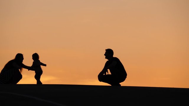 Little baby girl toddle from father to mother, telephoto perspective. Young family silhouettes against yellow sunset sky. Learn to walk or first steps concept. Framing with copyspace area at top right