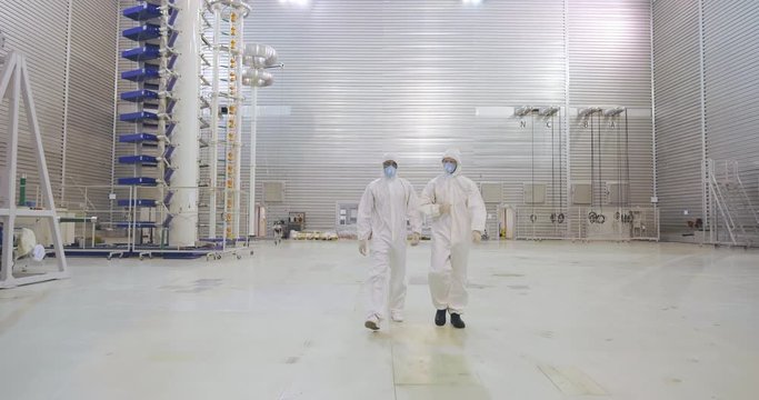 Two scientists in protective clothes finished experiment and go out of large testing laboratory throught huge opened doors