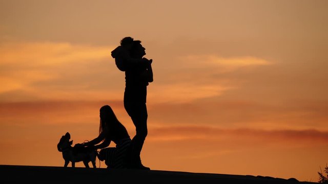 Young family silhouetted, woman hold and stir barking dog, man turn around on place with baby sitting on his shoulders. Orange sunset sky on backdrop. Copyspace version, empty area at on right half