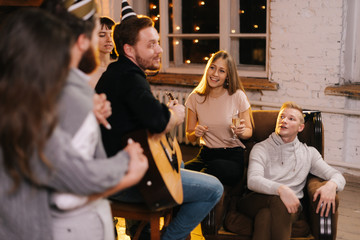 Happy young people are singing at the New Year party in cozy house. Happy young man plays guitar.