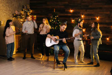 Group of young man and woman sings at a Christmas party in a cozy house. Happy young man plays...