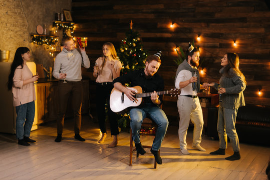Group of young man and woman sings at a Christmas party in a cozy house. Happy young man plays guitar. Christmas tree with garland and wall with festive illumination in background.