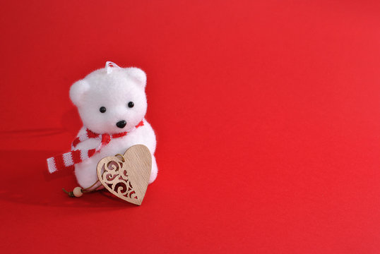 White Christmas decoration toy bear with striped scarf holding wooden design heart symbol decor on the red background with free blank copy text space in the right. Left oriented picture greeting card