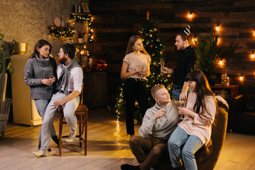Obraz premium Three young couples are talking and celebrating Christmas or new year at cozy home. Christmas tree with garland and wooden wall with festive illumination in background.